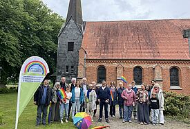 Eine Menschengruppe mit Regenbogenflaggen und einer Beachflag steht vor der Kirche in dem Dorf Hütten