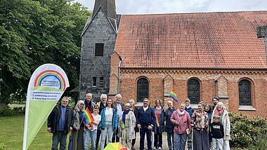 Eine Menschengruppe mit Regenbogenflaggen und einer Beachflag steht vor der Kirche in dem Dorf Hütten