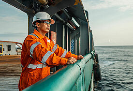 Ein Seemann in Arbeitskleidung lehnt an der Reling und schaut auf das offene Meer.
