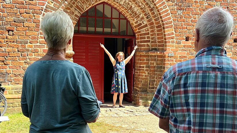 Maria Pulkenat vor Kirchenportal der Dorfkirche Poel Im Vordergrund stehen zwei ältere Teilnehmer einer Kirchenerkundung auf der Insel Poel. Sie blicken auf die Kirchenpädagogin Maria Pulkenat, die vor dem breiten und hohen Kirchenportal mit ausgebreiteten Armen steht.