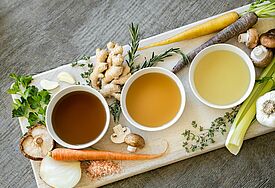 Auf dem Bild zu sehen sind drei kleine Schälchen mit Brühe in drei unterschiedlichen Farben. Drumherum verteilt liegen Zutaten für Brühe wie Zwiebel, Ingwer, Champignons. 