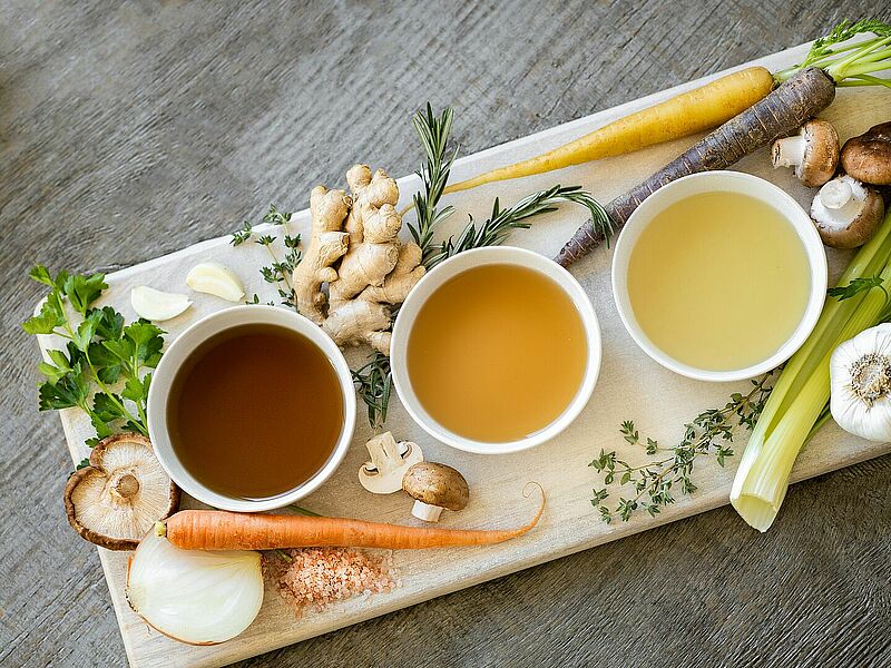 Fastensuppe Auf dem Bild zu sehen sind drei kleine Schälchen mit Brühe in drei unterschiedlichen Farben. Drumherum verteilt liegen Zutaten für Brühe wie Zwiebel, Ingwer, Champignons.