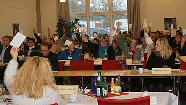 Menschen sitzen in Tischreihen und halten Zettel zur Abstimmung hoch