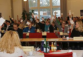 Menschen sitzen in Tischreihen und halten Zettel zur Abstimmung hoch
