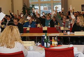 Menschen sitzen in Tischreihen und halten Zettel zur Abstimmung hoch
