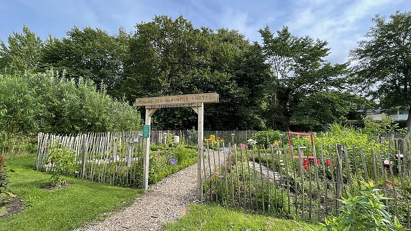 Ein Staktenzaun und ein Holztor führen in einen sommerlich blühenden kleinen Garten