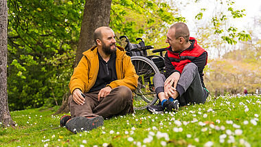 Rollstuhlfahrer und Freund sitzen auf einer Wiese. 