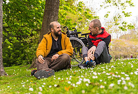 Rollstuhlfahrer und Freund sitzen auf einer Wiese. 