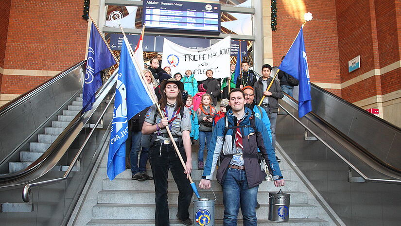 Eine Gruppe Pfadfinder läuft die Treppe vor dem Kieler Bahnhof herunter. Sie tragen die blaue Flagge mit dem Friedenslicht-Emblem und das Licht in geschützter Verpackung.