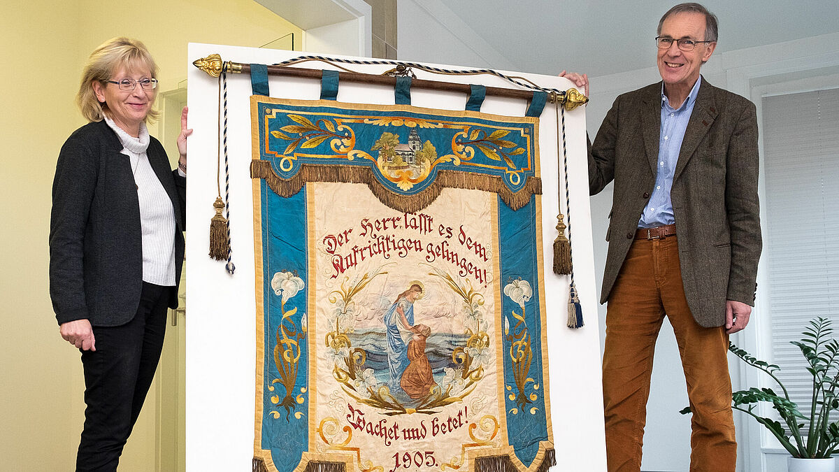 Archivleiterin Sybille Radtke-Kaak und Propst Stefan Block zeigen die restaurierte Fahne des kirchlichen Blau-Kreuz-Vereines in Neumünster.