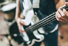Ein junger Mann spielt auf einer E-Gitarre, im Hintergrund sind weitere Instrumente wie eine Gitarre und ein Schlagzeug zu sehen. 