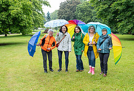 Fünf bunt gekleidete Frauen stehen mit aufgespannten Regenschirmen nebeneinander auf einer grünen Wiese.