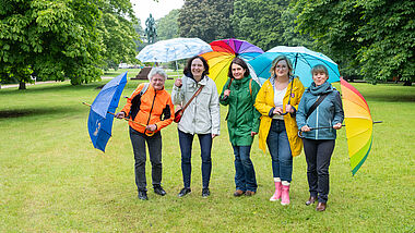 Fünf bunt gekleidete Frauen stehen mit aufgespannten Regenschirmen nebeneinander auf einer grünen Wiese.