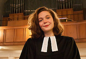 Vikarin Anna Böllert steht im Talar im Kirchenschiff, im Hintergrund ist die Orgel zu sehen.