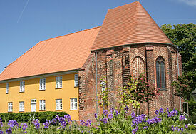 Das Bibelzentrum in Barth ist in einem gelben Gebäude untergebracht, davor stehen zwei Besucher, im Vordergrund blühen lila Blumen.