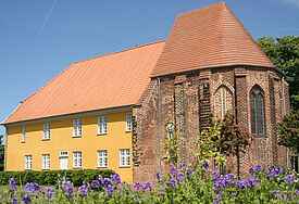 Das Gebäude des Barther Bibelzentrums leuchtet gelb in der Sommersonne, im Vordergrund blühen lila Blumen