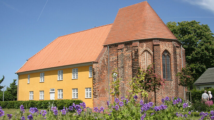 Das Gebäude des Barther Bibelzentrums leuchtet gelb in der Sommersonne, im Vordergrund blühen lila Blumen