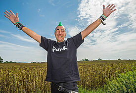 Mann mit grüner Irokesen-Frisur und Punk-T-Shirt reißt die Arme in die Höhe, dahinter ein Feld, Himmel blau, zartes Wolkenband
