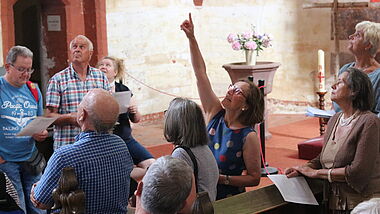 In einem mittelalterlichen Kirchenraum deutet Maria Pulkenat im bunten Kleid hoch zum Gewölbe. Interessierte Teilnehmer blicken ebenfalls hoch