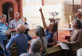 In einem mittelalterlichen Kirchenraum deutet Maria Pulkenat im bunten Kleid hoch zum Gewölbe. Interessierte Teilnehmer blicken ebenfalls hoch