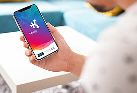 Man schaut einer Person über die Schulter, die ein Smartphone hält. Auf dem Display ist der Startbildschirm der Konapp in blau-pink zu sehen.