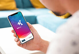 Man schaut einer Person über die Schulter, die ein Smartphone hält. Auf dem Display ist der Startbildschirm der Konapp in blau-pink zu sehen.