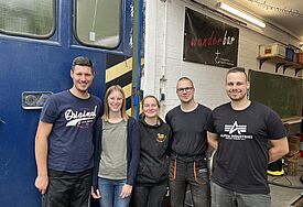 Fünf junge Männer und Frauen stehen nebeneinander und blicken fröhlich in die Kamera. Im Hintergrund ist ein blaues Werkstatttor und eine weiße Wand zu sehen. An der Wand hängt ein Plakat mit der Aufschrift "Wunderbar". 