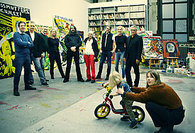 Gruppe von Menschen im Atelier, in dem sehr bunte Bilder hängen. Vorn ein Kind mit Roller, das von der Mutter festgehalten wird.