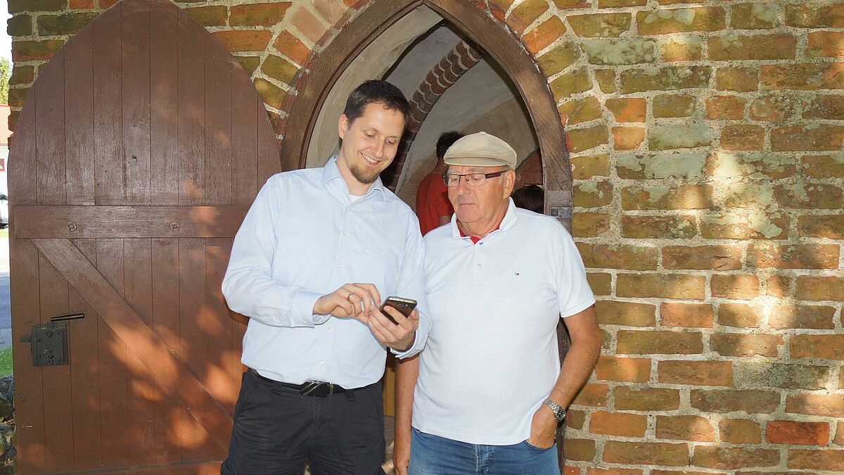 Vikar Friedemann Holmer (links) und Kirchengemeinderatsmitglied Klaus Gohlke überzeugen sich auf dem Smartphone während der Einweihung der "Erlebnis: Wallfahrt Levenhagen" von der ausgezeichneten Funktionsweise der App.