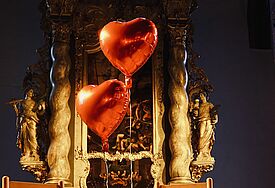Zwei Holzstühle mit roten schwebenden Herzballons stehen vor einem Altar.