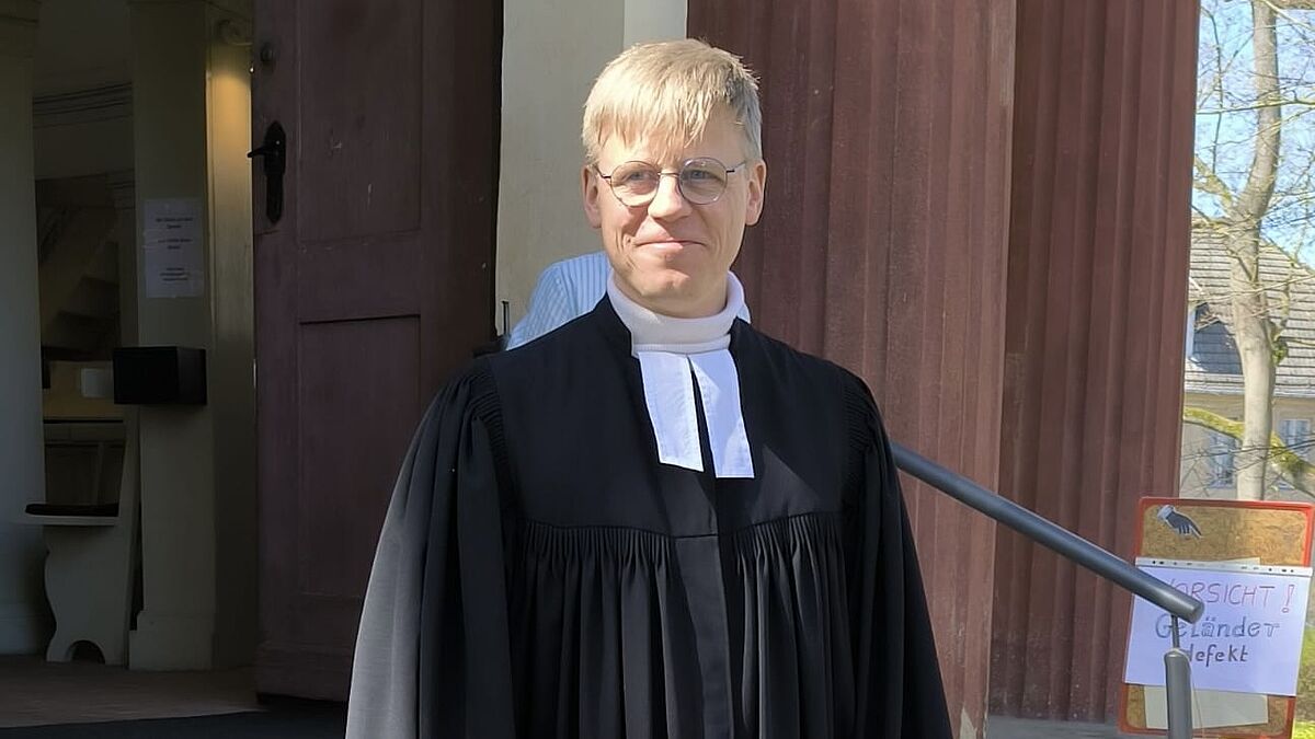 Pastor Nicolas Winkler in Talar vor einer Kirche.