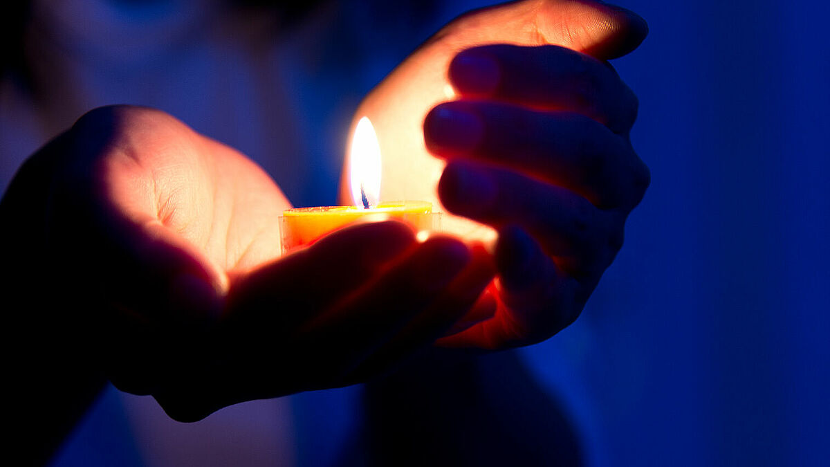 Ein Teelicht brennt gehalten in einer Hand, vor Wind geschützt von der zweiten Hand.
