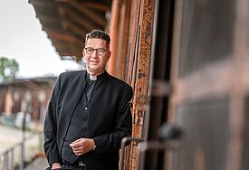Ein Mann mit kurzen Haaren und Brille lehnt seitlich an einer Wand. Er trägt ein schwarzes Collarhemd.