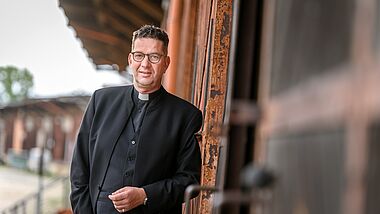 Ein Mann mit kurzen Haaren und Brille lehnt seitlich an einer Wand. Er trägt ein schwarzes Collarhemd.