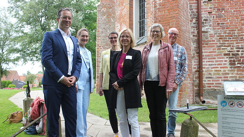 Sechs Menschen stehen vor einer alten Kirchenmauer aus Backstein und blicken in die Kamera