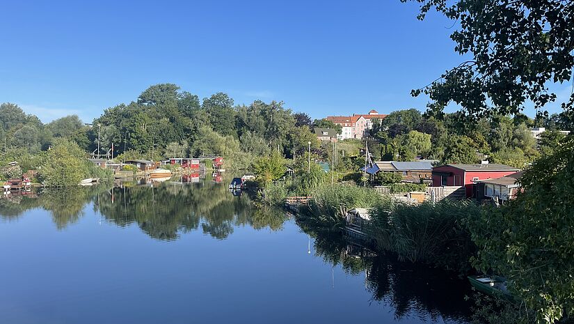 Der Blick über eine stille, blaue Wasserfläche. Im Hintergrund Gärten und Bootsstege. 