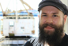 Mann mit Bart, Käppi auf dem Kopf, im Hintergrund verschwommen der Hafen