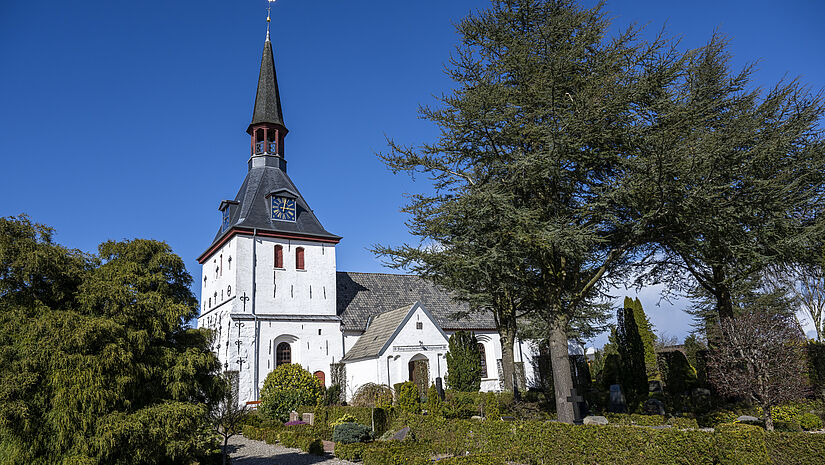 Blick auf eine weiße Kirche mit spitzem Turmhelm auf einem viereckigen Turm. 