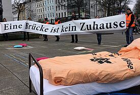 Menschen halten ein Banner in der Hand auf dem steht "Eine Brücke ist kein Zuhause", im Vordergrund steht ein Bett mit einer orangen Decke auf der Straße.
