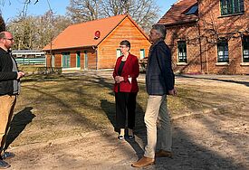 Landesbischöfin Kristina Kühnbaum-Schmidt (Mitte) steht mit dem Leiter der Unterkunft in Damm, Michael Güldner (links), und dem Landeskirchlichen Beauftragten Mecklenburg-Vorpommern Markus Wiechert zusammen auf einer Rasenfläche vor mehreren Häusern.