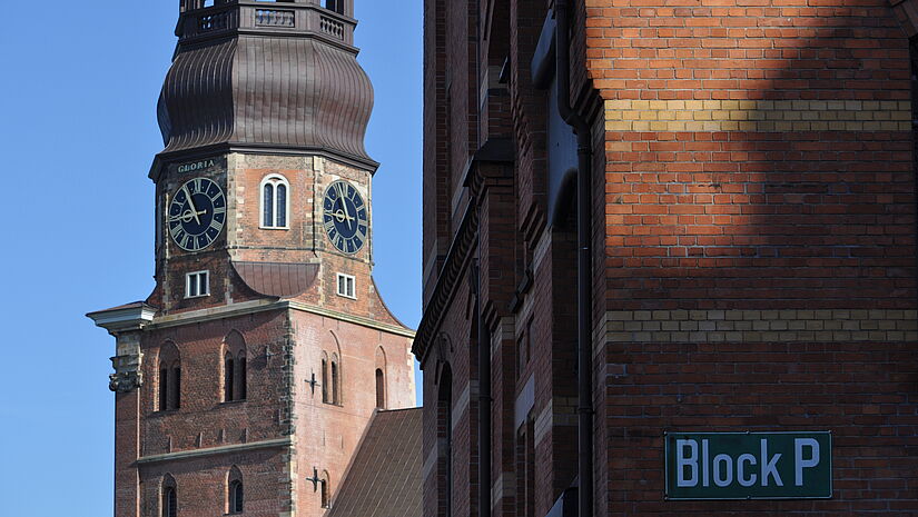 Backsteinkirche, angeschnitten, man sieht den Turm mit Ziffernblatt, etwas vom Dach, rechts ist die Wand eines anderen Gebäudes zu sehen. Der himmel ist blau, die Sonne scheint