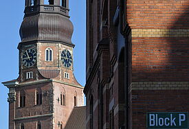 Backsteinkirche, angeschnitten, man sieht den Turm mit Ziffernblatt, etwas vom Dach, rechts ist die Wand eines anderen Gebäudes zu sehen. Der himmel ist blau, die Sonne scheint