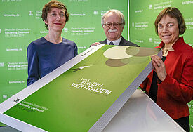 Die drei Menschen halten das Programm als grünes Riesenbuch in den Händen und lächeln imn die kamera