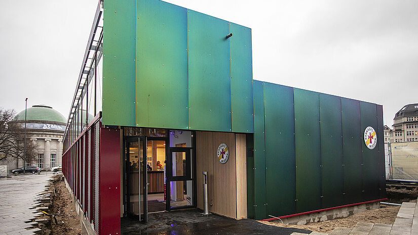 Im Neubau der Bahnhofsmission Hamburg ist jeder willkommen. Der Neubau aus Holz schimmert grünlich-blau.
