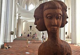 Im Vordergrund ist die Schnitzerei einer Figur, die mehrere Gesichter trägt, im Hintergrund ist der Kirchraum von St. Petri zu sehen, der mit weißen Stoffbahnen behängt ist.