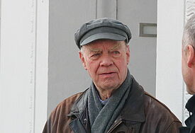 Heiko Lietz steht mit brauner Jacke und grauer Mütze vor einer Hauswand. Er blickt zu Bischof Jeremias, der von hinten am rechten Bildrand zu erkennen ist.