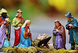 Bunte Krippenfiguren von Maria und Josef ums Jesuskind kniend eingerahmt von den Heiligen drei Königen und einem Hirten stehen vor einem bunten Hintergrund