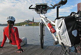 Eine Frau mit Fahrradhelm sitzt auf einem Steg vor dem See, daneben steht ihr Fahrrad mit Fahrradtaschen, im Hintergrund ist über den See hinweg die Kirche zu sehen
