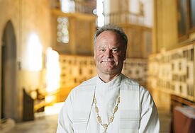 Bischof Tilman Jeremias steht in einer weißen Albe und weißer Stola im goldenen Licht des Domfensters des Greifswalder Doms St. Nikolai