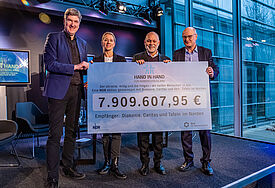 Drei Männer und eine Frau halten einen symbolischen, übergrossen Scheck in der Hand. 7.909.607,95 Euro steht darauf geschrieben.